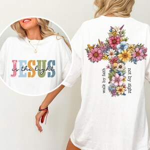 May include: White t-shirt with the text "JESUS is the light" in colorful letters on the front. The back features a floral cross design with the words "walk by faith not by sight".