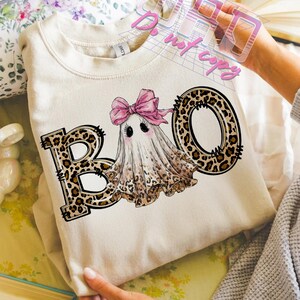 May include: Cream-colored sweatshirt with a Halloween-themed graphic. The graphic features the word "BOO" with leopard print letters, and a cute ghost wearing a pink bow. The ghost has black eyes and blush.