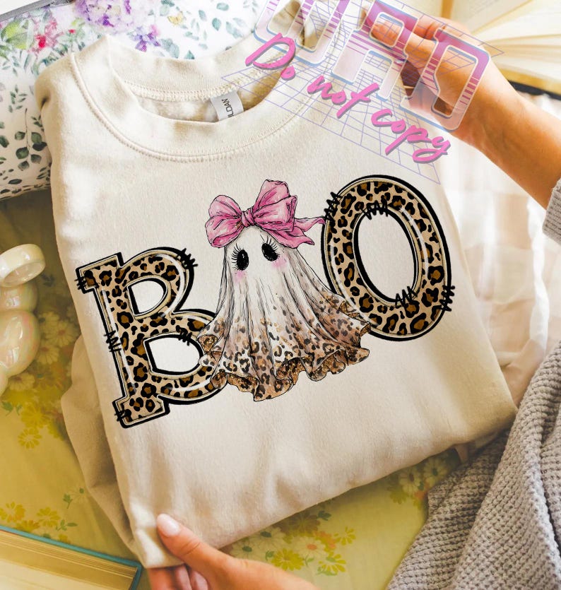 May include: Cream-colored sweatshirt with a Halloween-themed graphic. The graphic features the word "BOO" with leopard print letters, and a cute ghost wearing a pink bow. The ghost has black eyes and blush.