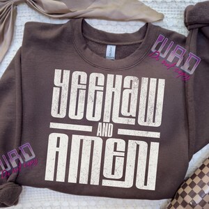 May include: A brown sweatshirt with the words "YEEHAW AND AMEN" printed in a distressed white font. The sweatshirt has a crew neck and long sleeves. The text is centered on the front of the garment.