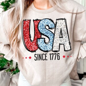 May include: A beige sweatshirt with a glittery red, white, and blue "USA" graphic and the text "SINCE 1776" in black.