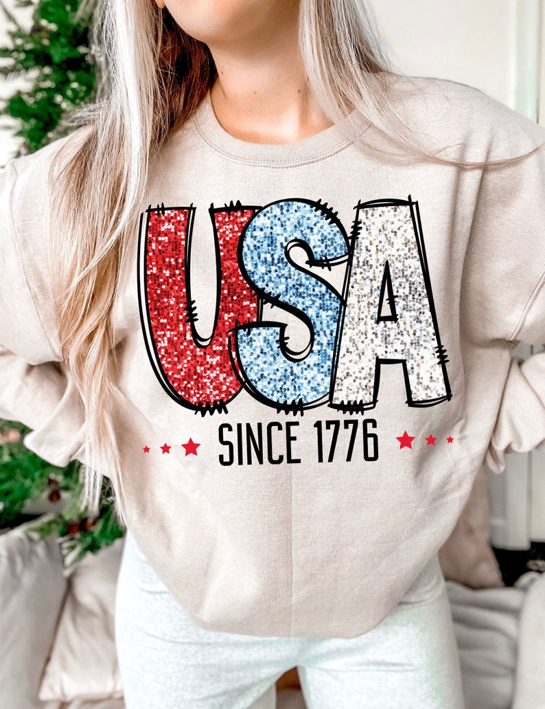 May include: A beige sweatshirt with a glittery red, white, and blue "USA" graphic and the text "SINCE 1776" in black.