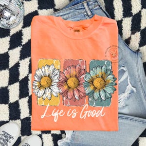 May include: Coral-colored t-shirt featuring a floral design with three daisies in yellow, pink, and blue rectangular frames. The phrase "Life is Good" is printed in white script below the flowers. The shirt is folded on a denim surface.