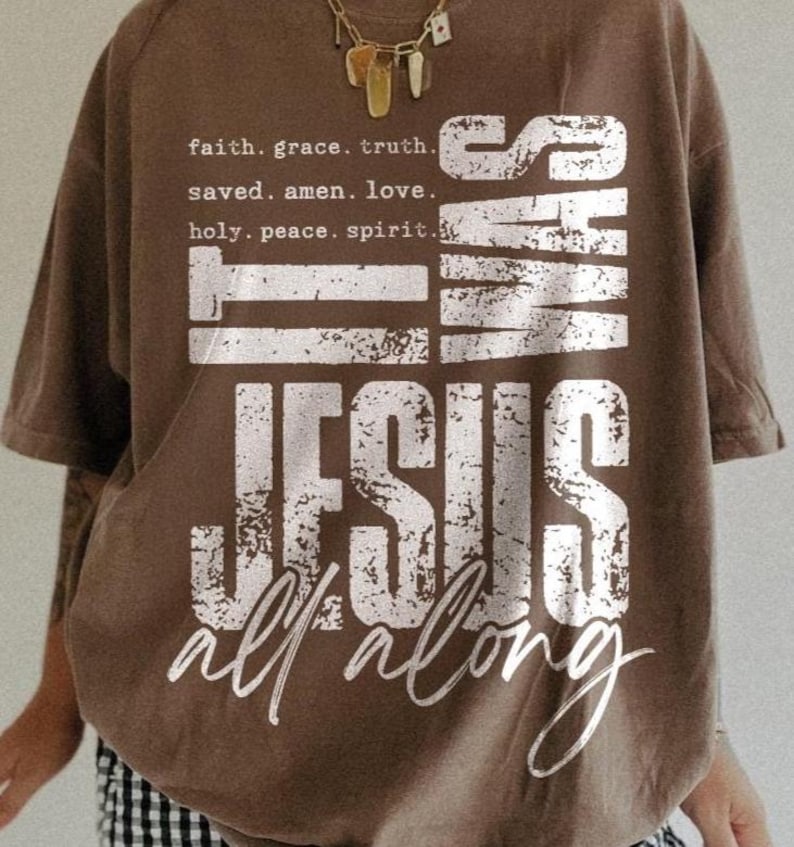May include: Brown t-shirt with the text "WAS JESUS all along" in distressed white lettering. The shirt also features the phrases "faith. grace. truth.", "saved. amen. love.", and "holy. peace. spirit." A gold necklace is visible.