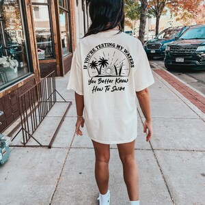 May include: A white t-shirt with a black graphic that says "If you're testing my waters you better know how to swim". The graphic features palm trees, a sun, and surfboards.
