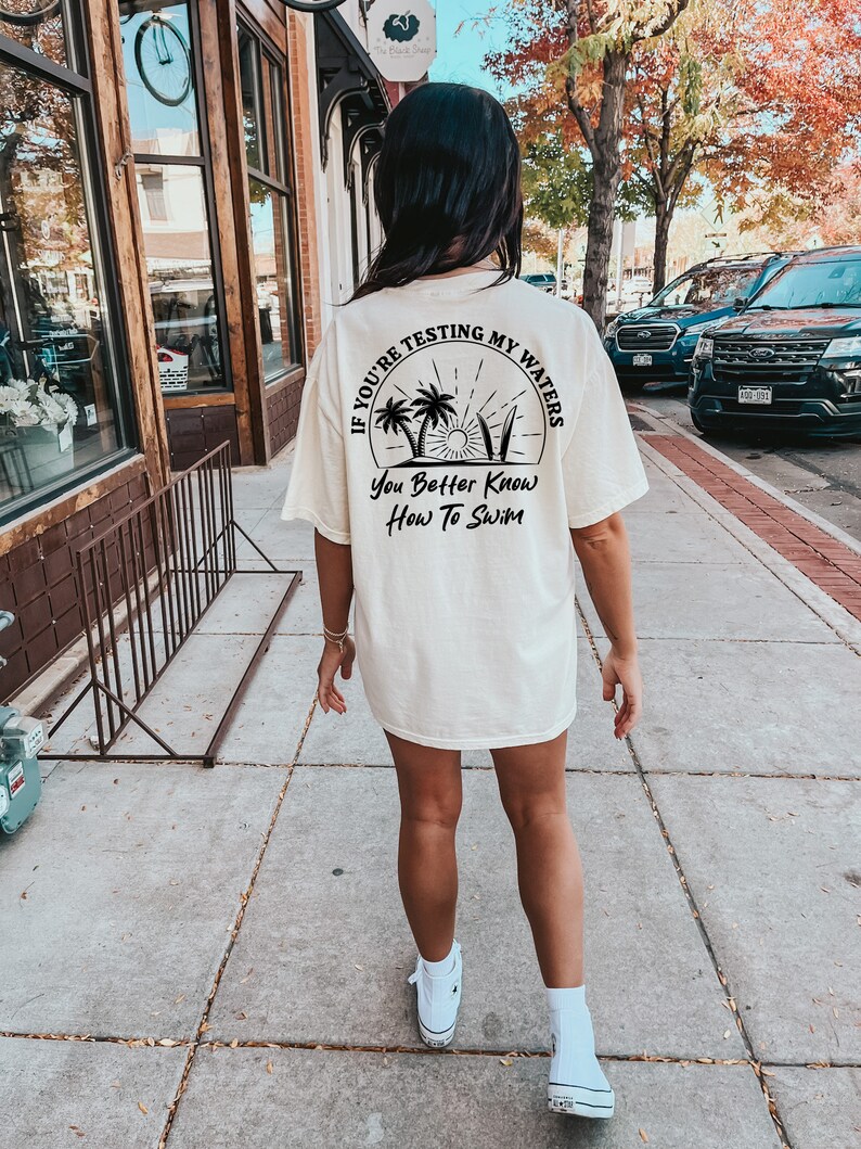 May include: A white t-shirt with a black graphic that says "If you're testing my waters you better know how to swim". The graphic features palm trees, a sun, and surfboards.