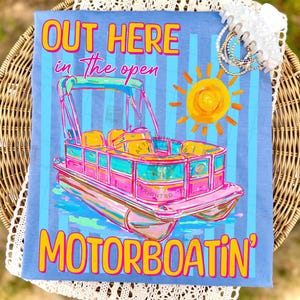 May include: A light blue t-shirt with a colorful illustration of a pontoon boat, a yellow sun, and the text "OUT HERE in the open MOTORBOATIN'". The boat is pink, teal, and yellow.