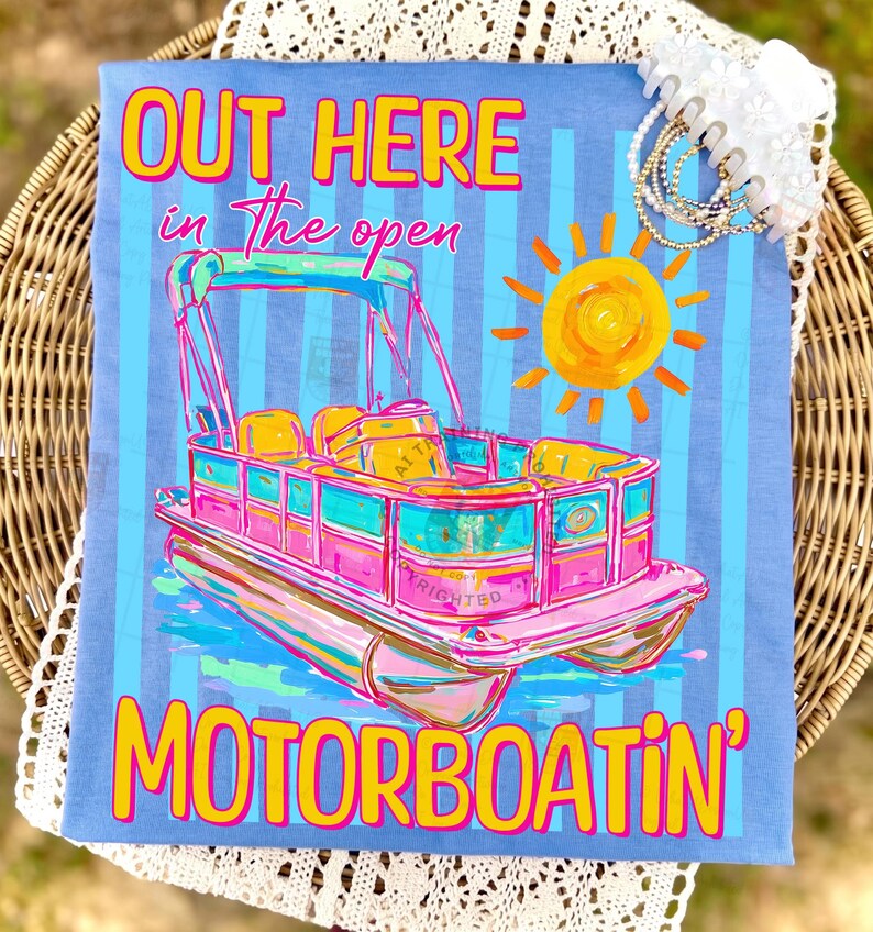 May include: A light blue t-shirt with a colorful illustration of a pontoon boat, a yellow sun, and the text "OUT HERE in the open MOTORBOATIN'". The boat is pink, teal, and yellow.