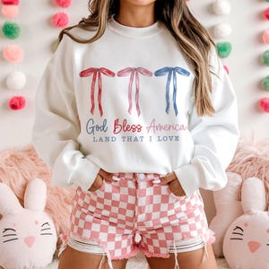 May include: White sweatshirt with a red, white and blue bow design and the text "God Bless America Land That I Love".