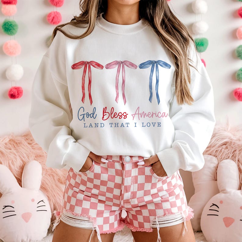May include: White sweatshirt with a red, white and blue bow design and the text "God Bless America Land That I Love".