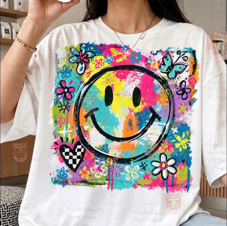 May include: White t-shirt featuring a colorful smiley face design. The graphic includes a black smiley face, flowers, a butterfly, and a checkered heart, all set against a vibrant, multi-colored background. The text "WHAT A DESIGN" is visible.