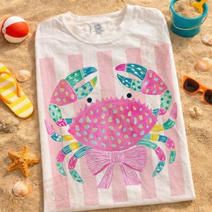 May include: White t-shirt with a pink crab design. The crab has a pink body with colorful accents and a pink bow. The background features pink and white stripes. Beach-themed accessories like a beach ball, sunglasses, and seashells are scattered around the shirt.