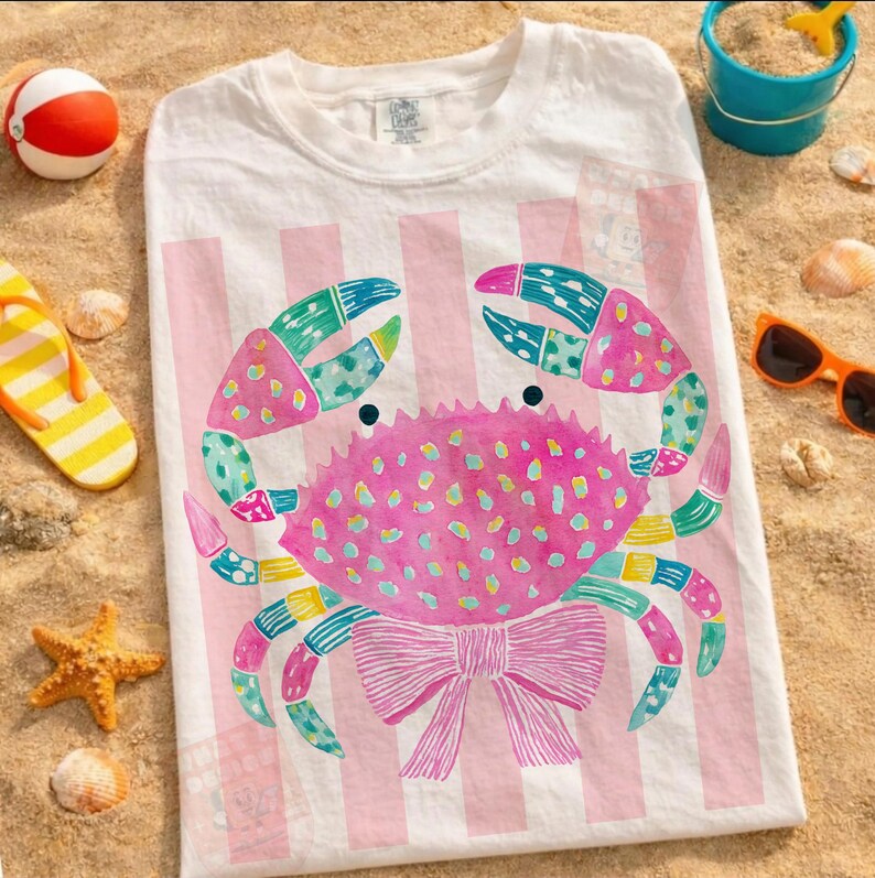 May include: White t-shirt with a pink crab design. The crab has a pink body with colorful accents and a pink bow. The background features pink and white stripes. Beach-themed accessories like a beach ball, sunglasses, and seashells are scattered around the shirt.