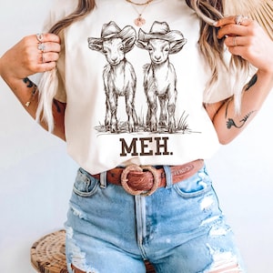 May include: White t-shirt with a graphic of two goats wearing cowboy hats. The text "MEH." is printed below the goats.