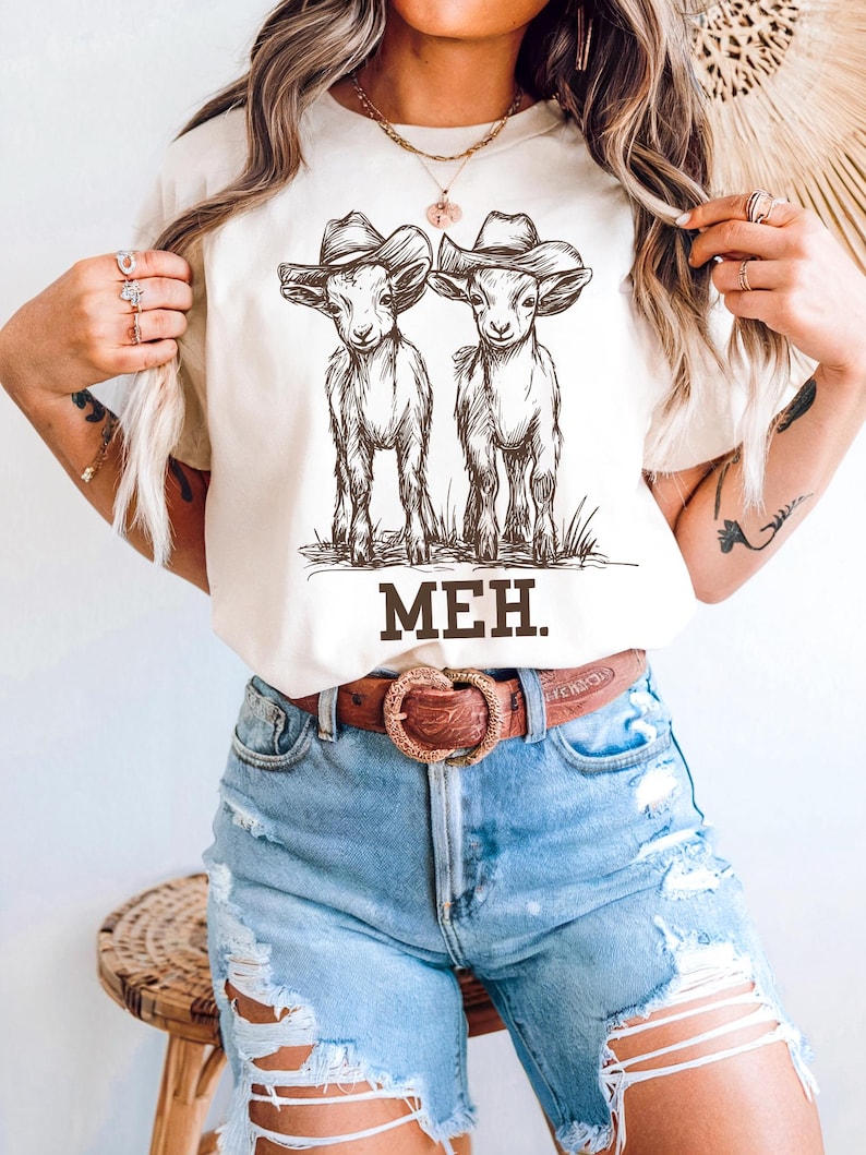 May include: White t-shirt with a graphic of two goats wearing cowboy hats. The text "MEH." is printed below the goats.