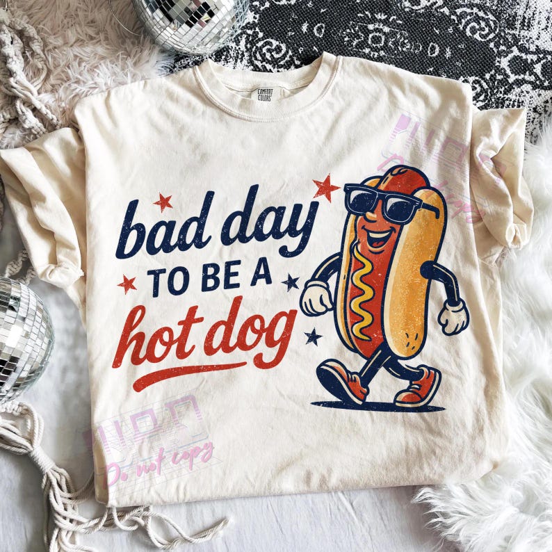 Puede incluir: Camiseta color crema con la frase "bad day to be a hot dog" en azul marino y rojo. Un dibujo animado de un perrito caliente con gafas de sol y zapatillas deportivas est&aacute; impreso en la camiseta.