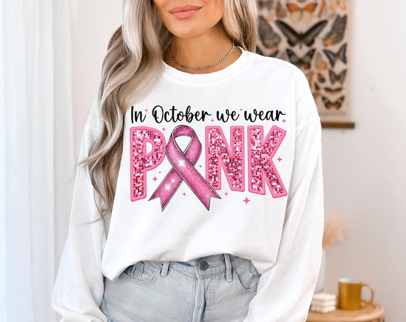 May include: White sweatshirt with long sleeves and a pink glitter design that says "In October we wear PINK" with a pink ribbon.