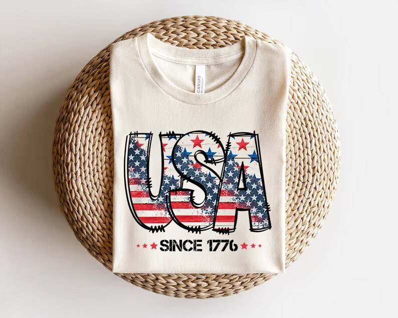 May include: A white t-shirt with a distressed graphic of the word "USA" in red, white, and blue with stars. The graphic is surrounded by stars and the text "Since 1776" is below the graphic.