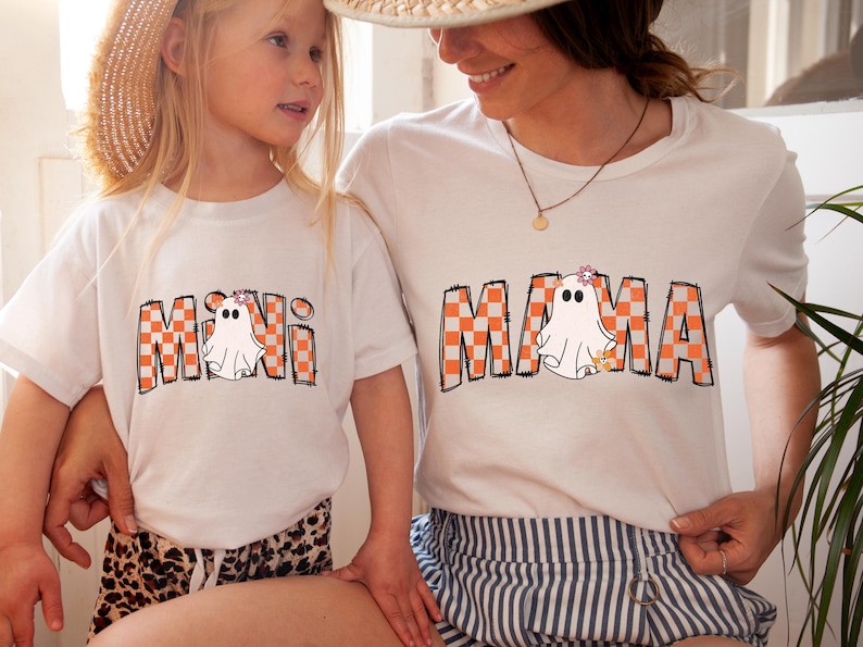Pu&ograve; includere: Due magliette bianche con una grafica di un fantasma e un testo a scacchi. La maglietta pi&ugrave; grande dice "MAMA" e la maglietta pi&ugrave; piccola dice "MINI".