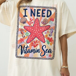 May include: Cream-colored t-shirt with a colorful graphic. The graphic features a red starfish, seashells, seahorses, and the text "I NEED Vitamin Sea" in a decorative frame. The design has a vintage, beachy vibe.