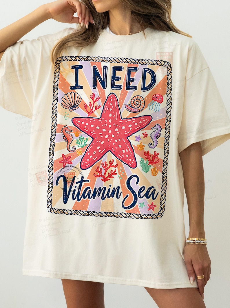 May include: Cream-colored t-shirt with a colorful graphic. The graphic features a red starfish, seashells, seahorses, and the text "I NEED Vitamin Sea" in a decorative frame. The design has a vintage, beachy vibe.