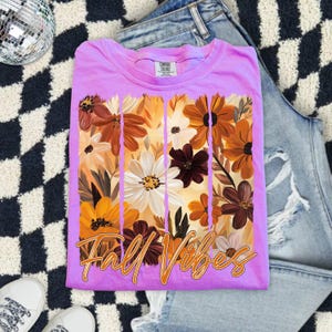 May include: A lilac-colored t-shirt with a floral design in shades of brown, orange, and white. The words "Fall Vibes" are written in an orange script across the bottom of the floral design. The shirt is folded on top of a pair of blue jeans.