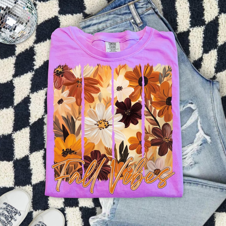 May include: A lilac-colored t-shirt with a floral design in shades of brown, orange, and white. The words "Fall Vibes" are written in an orange script across the bottom of the floral design. The shirt is folded on top of a pair of blue jeans.