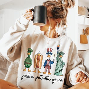 May include: Cream-colored sweatshirt with a graphic of four cartoon geese dressed as patriotic figures. The geese are a soldier, a hot dog, Uncle Sam, and the Statue of Liberty. The text "just a patriotic goose" is below the graphic.