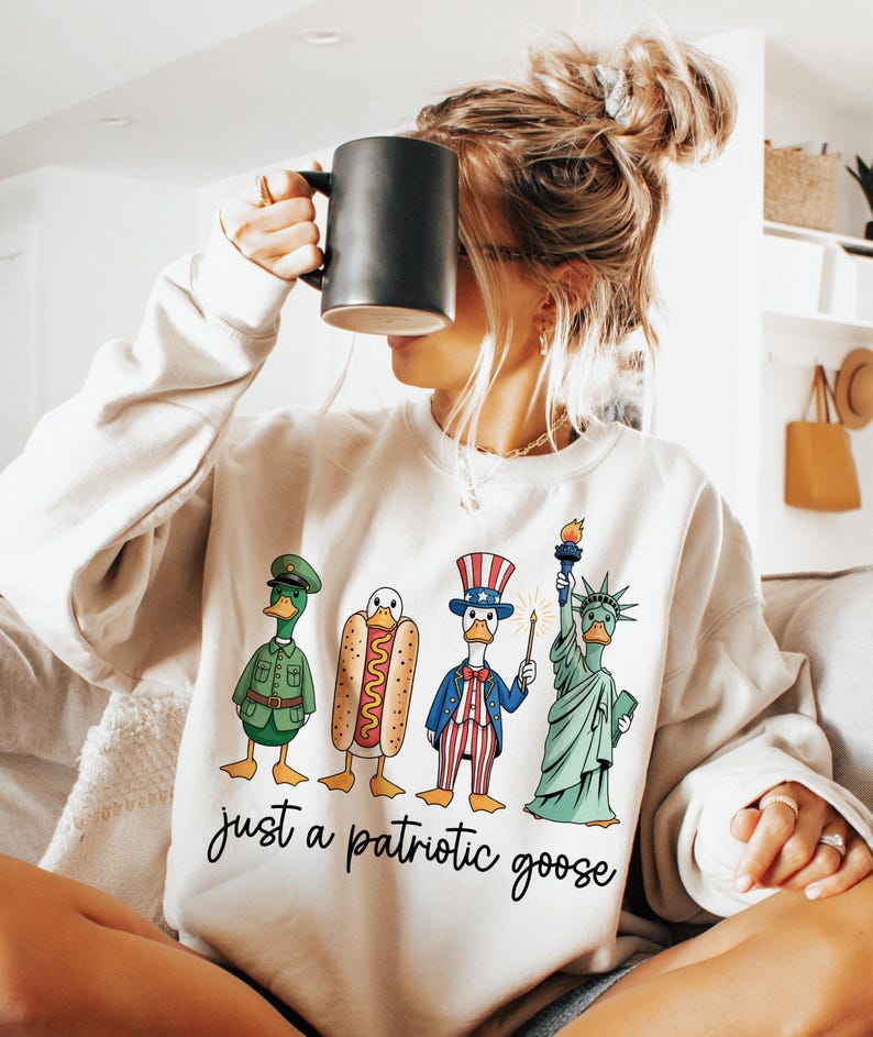 May include: Cream-colored sweatshirt with a graphic of four cartoon geese dressed as patriotic figures. The geese are a soldier, a hot dog, Uncle Sam, and the Statue of Liberty. The text "just a patriotic goose" is below the graphic.