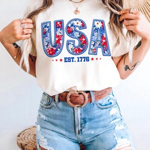 May include: Cream-colored t-shirt with the word "USA" in blue, red, and white floral design. The text "EST. 1776" is below the word "USA" with red stars on each side. The shirt is paired with light blue denim shorts and a brown belt.