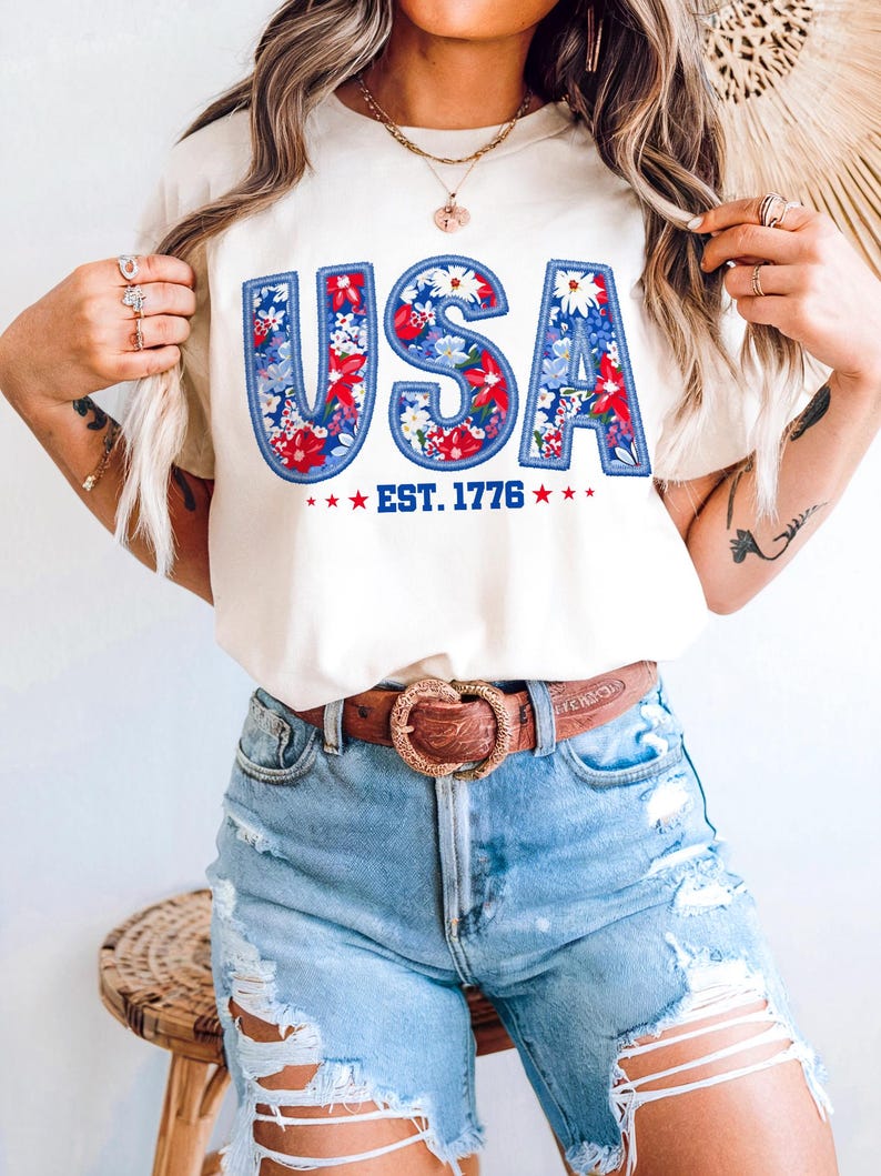 May include: Cream-colored t-shirt with the word "USA" in blue, red, and white floral design. The text "EST. 1776" is below the word "USA" with red stars on each side. The shirt is paired with light blue denim shorts and a brown belt.