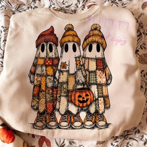 May include: Cream-colored sweatshirt featuring a graphic of three cartoon ghosts in patchwork outfits and orange beanies. The ghosts are holding a jack-o'-lantern bucket and wearing orange sneakers. A fun, festive design for Halloween.