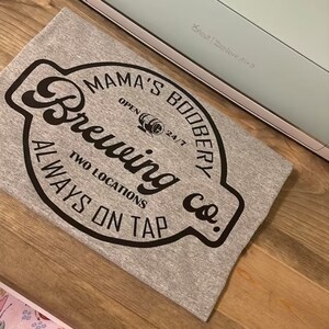 May include: A gray t-shirt with a black circular design that says "Mama's Boobery Brewing Co." with the text "Always on Tap" and "Two Locations" below the logo.