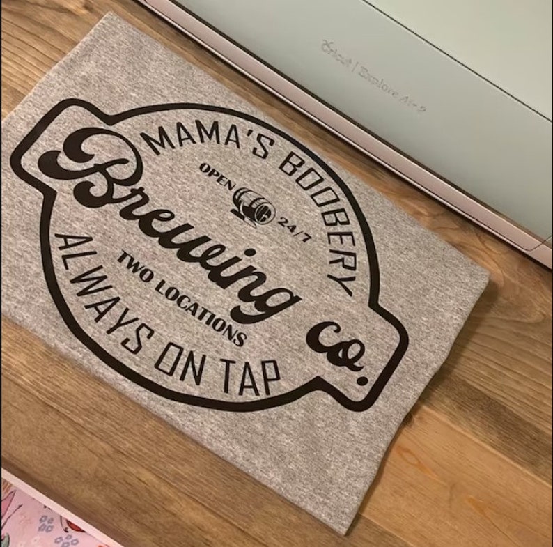 May include: A gray t-shirt with a black circular design that says "Mama's Boobery Brewing Co." with the text "Always on Tap" and "Two Locations" below the logo.