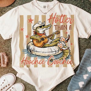May include: Cream-colored t-shirt featuring a cartoon alligator wearing a cowboy hat and sunglasses, playing a guitar while floating on an inner tube. The text "Hotter Than A Hoochie Coochie" is printed on the shirt.