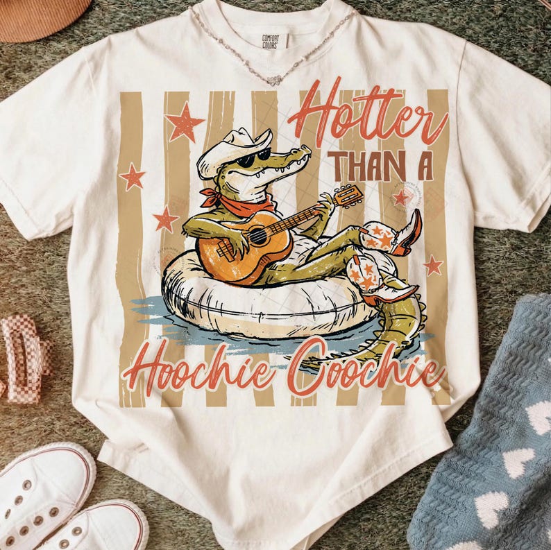 May include: Cream-colored t-shirt featuring a cartoon alligator wearing a cowboy hat and sunglasses, playing a guitar while floating on an inner tube. The text "Hotter Than A Hoochie Coochie" is printed on the shirt.