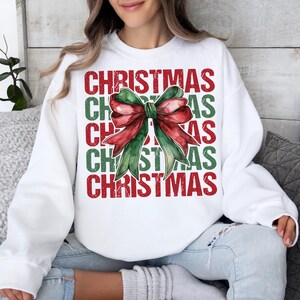 May include: White Christmas sweatshirt featuring a watercolor bow in red and green. The word "CHRISTMAS" is stacked vertically in red and green letters. The sweatshirt is a festive holiday apparel item.