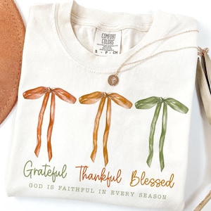 May include: A cream-coloured t-shirt with three watercolour bows in orange and green. The words "Grateful, Thankful, Blessed" are printed below the bows, with the phrase "God is faithful in every season" underneath. A gold necklace rests on the shirt.