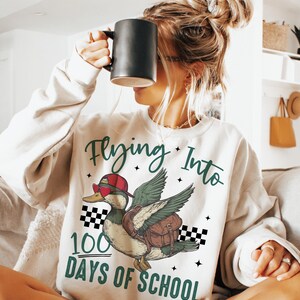 May include: White sweatshirt with a green and black graphic of a duck wearing sunglasses and a hat, flying with a backpack. The text "Flying Into 100 Days of School" is printed on the sweatshirt.
