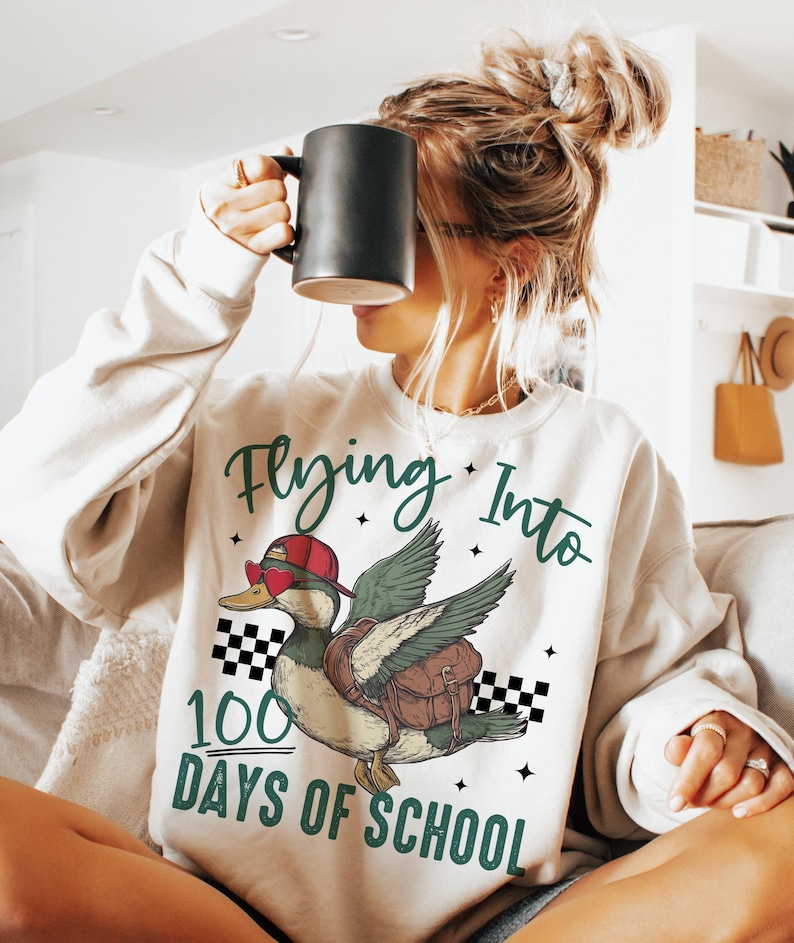 May include: White sweatshirt with a green and black graphic of a duck wearing sunglasses and a hat, flying with a backpack. The text "Flying Into 100 Days of School" is printed on the sweatshirt.