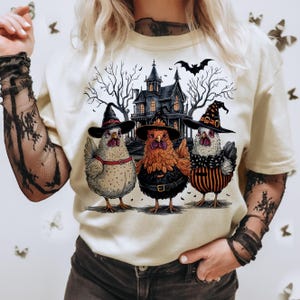 K&ouml;nnte beinhalten: Ein wei&szlig;es T-Shirt mit einem Aufdruck von drei H&uuml;hnern, die Hexenh&uuml;te tragen und vor einem Spukhaus stehen. Die H&uuml;hner sind wei&szlig;, orange und schwarz-wei&szlig;.