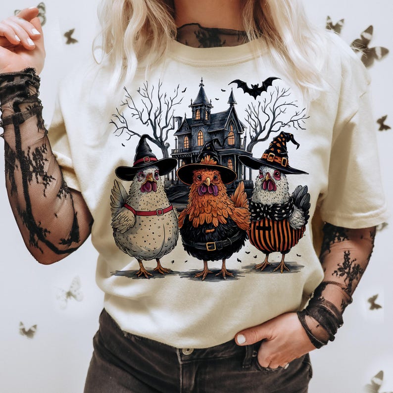 K&ouml;nnte beinhalten: Ein wei&szlig;es T-Shirt mit einem Aufdruck von drei H&uuml;hnern, die Hexenh&uuml;te tragen und vor einem Spukhaus stehen. Die H&uuml;hner sind wei&szlig;, orange und schwarz-wei&szlig;.