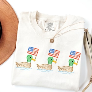May include: Cream-colored Comfort Colors t-shirt featuring three embroidered ducks, each holding a small American flag. The ducks are depicted in a cartoon style, with green heads, yellow beaks, and brown bodies. A gold necklace and a brown hat are also visible.