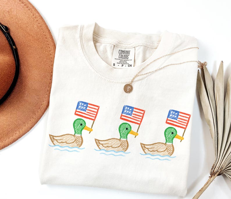May include: Cream-colored Comfort Colors t-shirt featuring three embroidered ducks, each holding a small American flag. The ducks are depicted in a cartoon style, with green heads, yellow beaks, and brown bodies. A gold necklace and a brown hat are also visible.