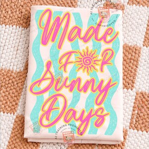 May include: A folded light pink t-shirt with the words "Made For Sunny Days" in bright pink script. The text is layered over wavy teal lines and a yellow sun graphic. The shirt is on a patterned beige and white surface.
