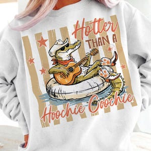 May include: White sweatshirt with a graphic of an alligator wearing a cowboy hat, sunglasses, and boots, playing a guitar while floating on an inner tube. Text reads "Hotter Than A Hoochie Coochie."