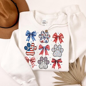 May include: White sweatshirt with a red, white, and blue paw print and bow design. The design features a variety of patterns, including stars, stripes, and polka dots.