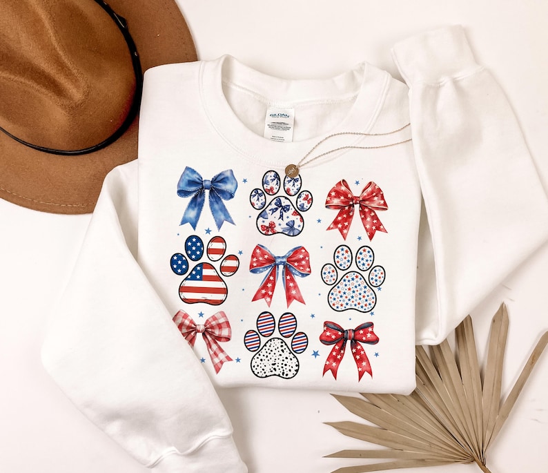 May include: White sweatshirt with a red, white, and blue paw print and bow design. The design features a variety of patterns, including stars, stripes, and polka dots.