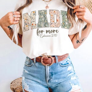 May include: Cream-colored t-shirt with the words "MADE for more" in a floral design. The text "Ephesians 2:10" is below the main text. The shirt is paired with blue denim shorts and a brown belt.