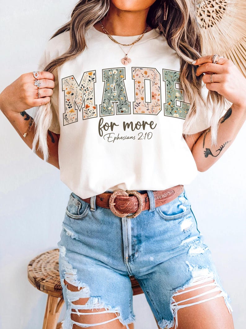 May include: Cream-colored t-shirt with the words "MADE for more" in a floral design. The text "Ephesians 2:10" is below the main text. The shirt is paired with blue denim shorts and a brown belt.